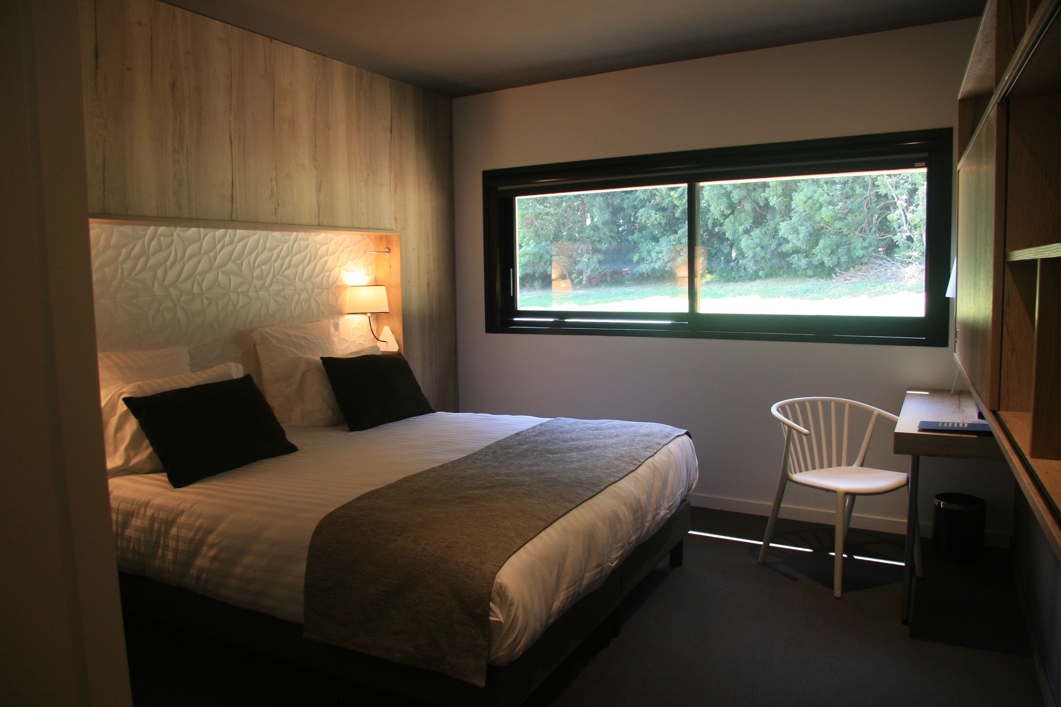 Een slaapkamer met een bed, een stoel, een bureau en een raam met uitzicht op bomen.
