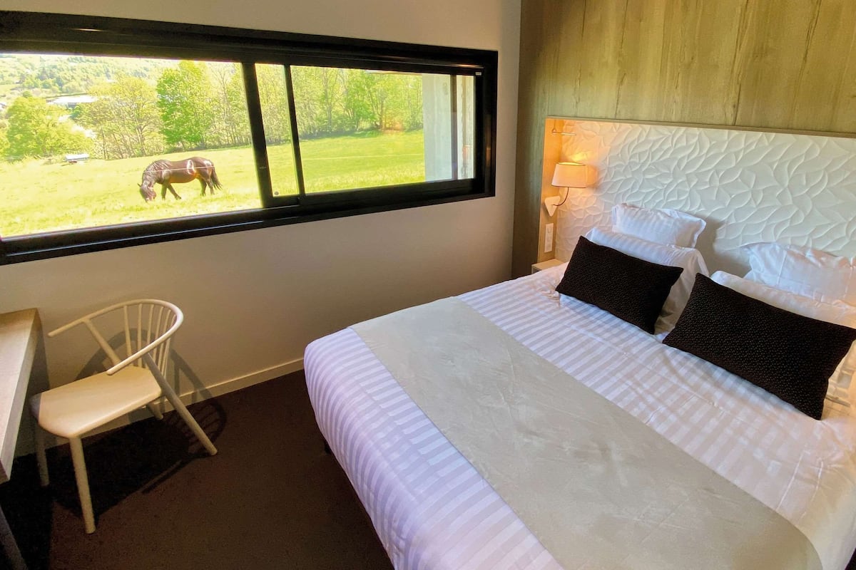 Een slaapkamer met een groot bed, een bureau, een stoel en een raam met uitzicht op een paard dat graast.