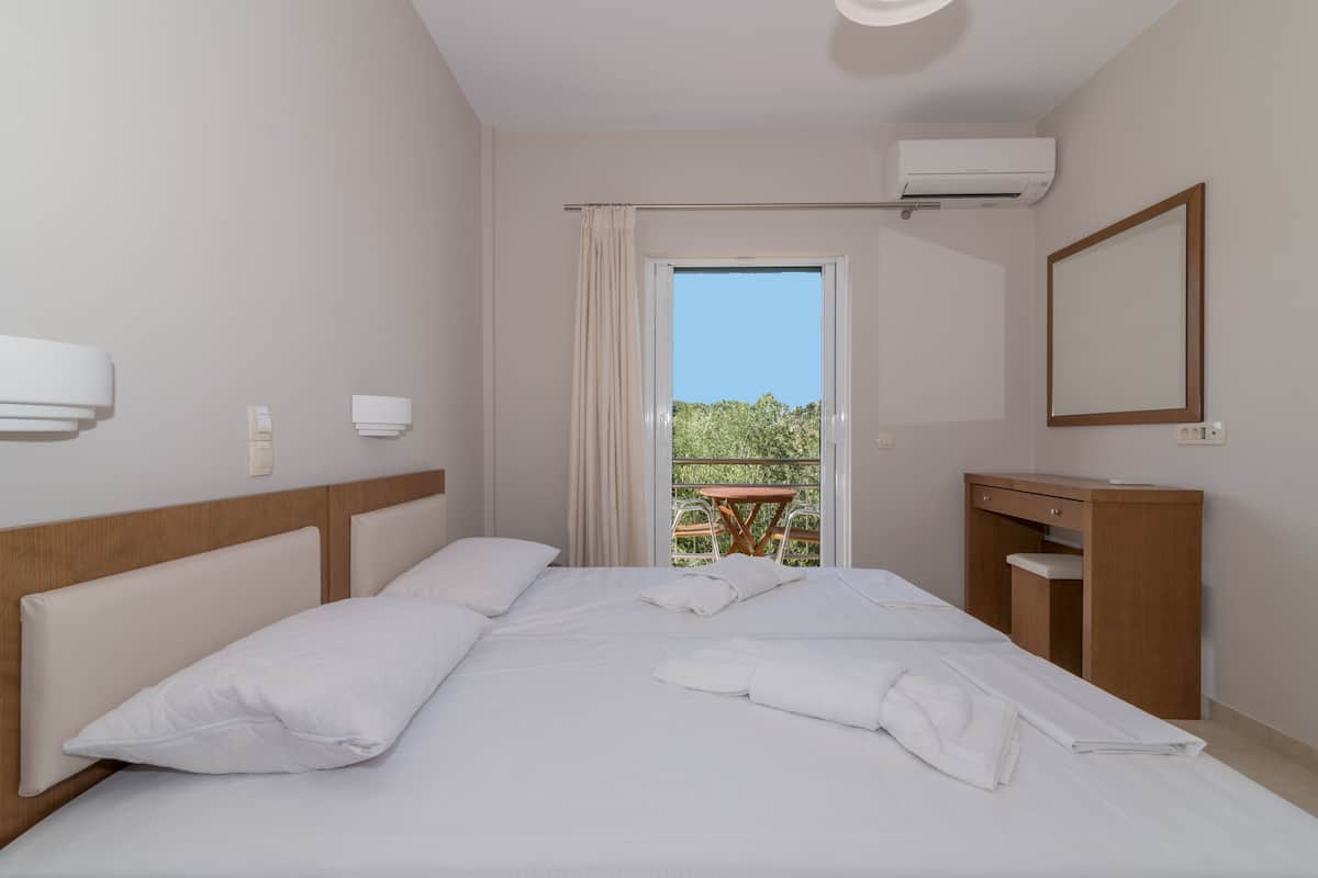 A hotel room with a large bed, a wooden nightstand, a mirror, and a view of greenery outside.