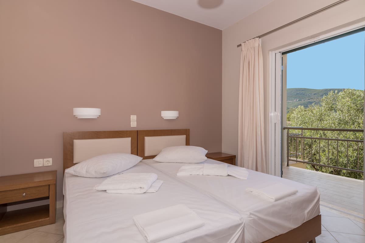 A neatly made bed with white linens and towels, a wooden headboard, bedside tables with wall-mounted lamps, and a balcony with a view of greenery.