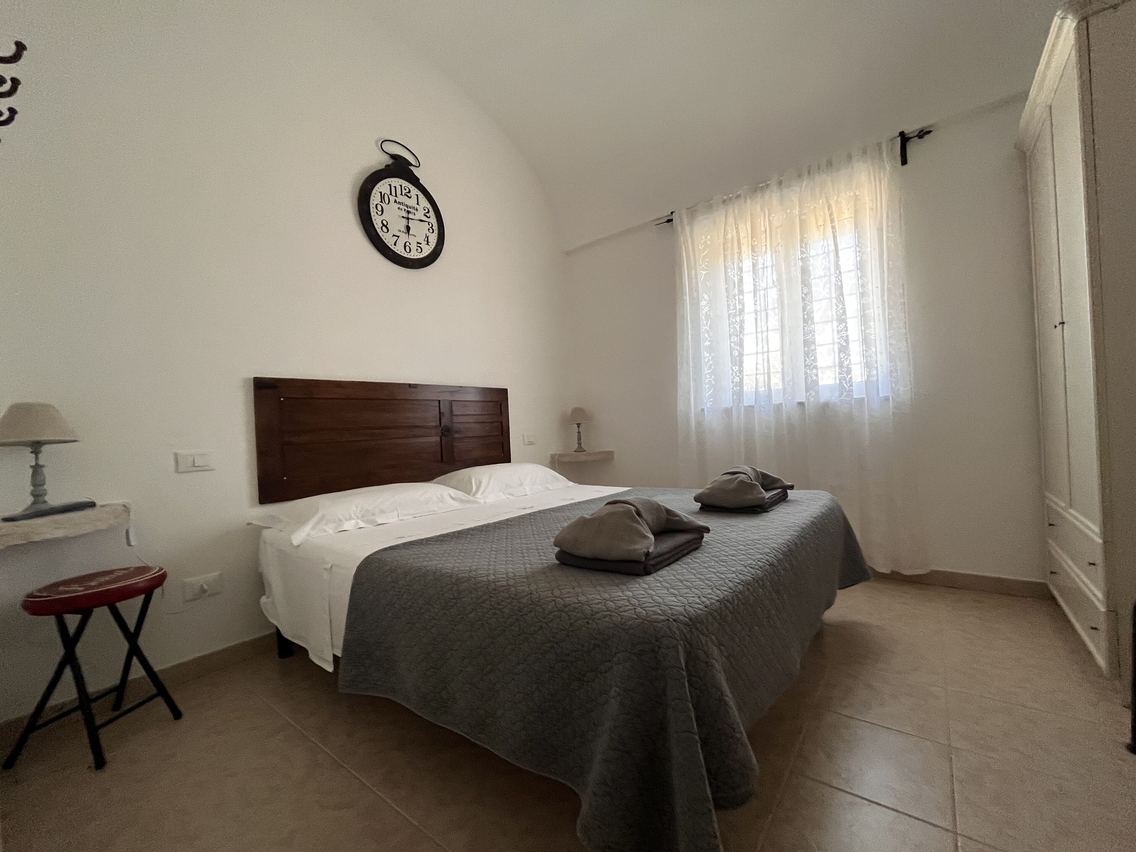 A bedroom with a wooden headboard, a bed with a grey quilt, a bedside table with a lamp, and a wall clock.