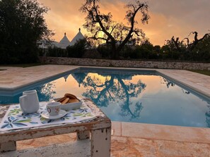 A swimming pool with a table and chairs in the foreground, a sunset in the background, and a stone wall surrounding the area. - "Al Guappo D'oro," trullo with pool nestled in the Apulian countryside.  (Ostuni)