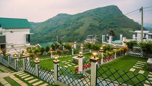 A landscaped garden with a pathway, a white building with a green roof, and a mountain in the background. - Palette Hill View Resort Vagamon (Peermade)