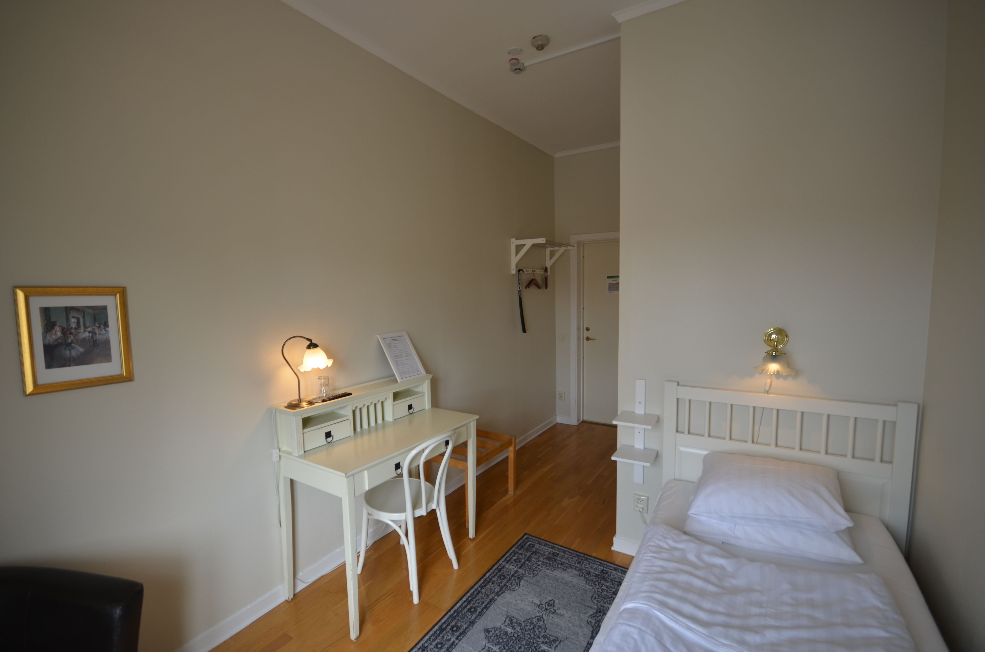 Un dormitorio con cama, escritorio, silla, un tapete pequeño y una lámpara fijada a la pared.