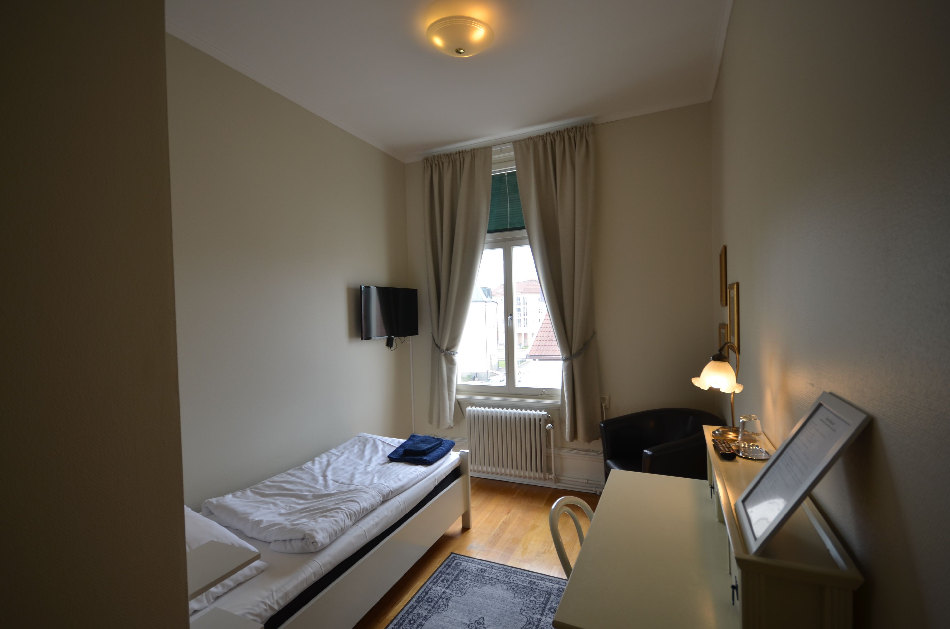 Une petite pièce bien éclairée, avec un lit simple, un bureau doté d’une lampe, une chaise et une fenêtre avec des rideaux.