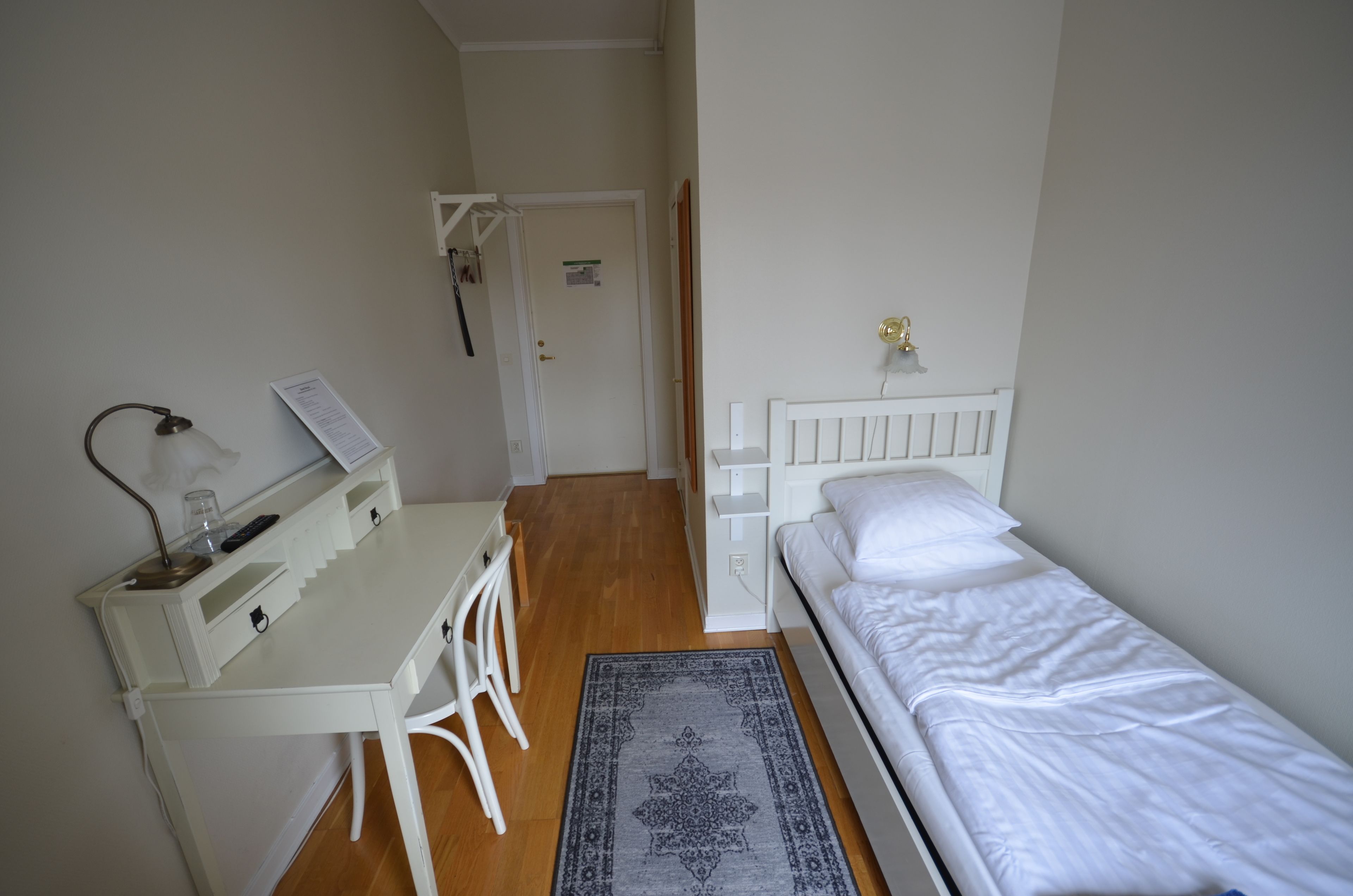 A small, clean room with a white desk, a chair, a bed, and a door leading to another room.