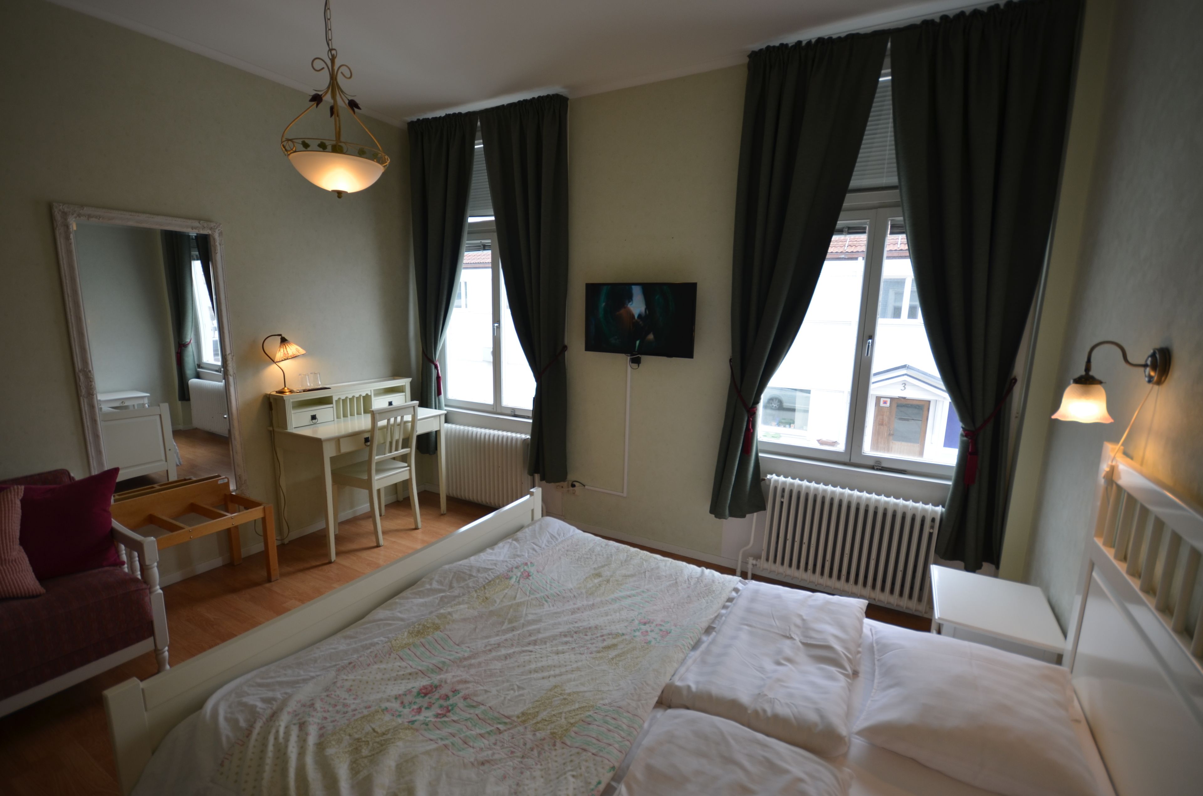 A bedroom with a bed, a desk, a chair, a dresser, a mirror, a television, and a window with curtains.