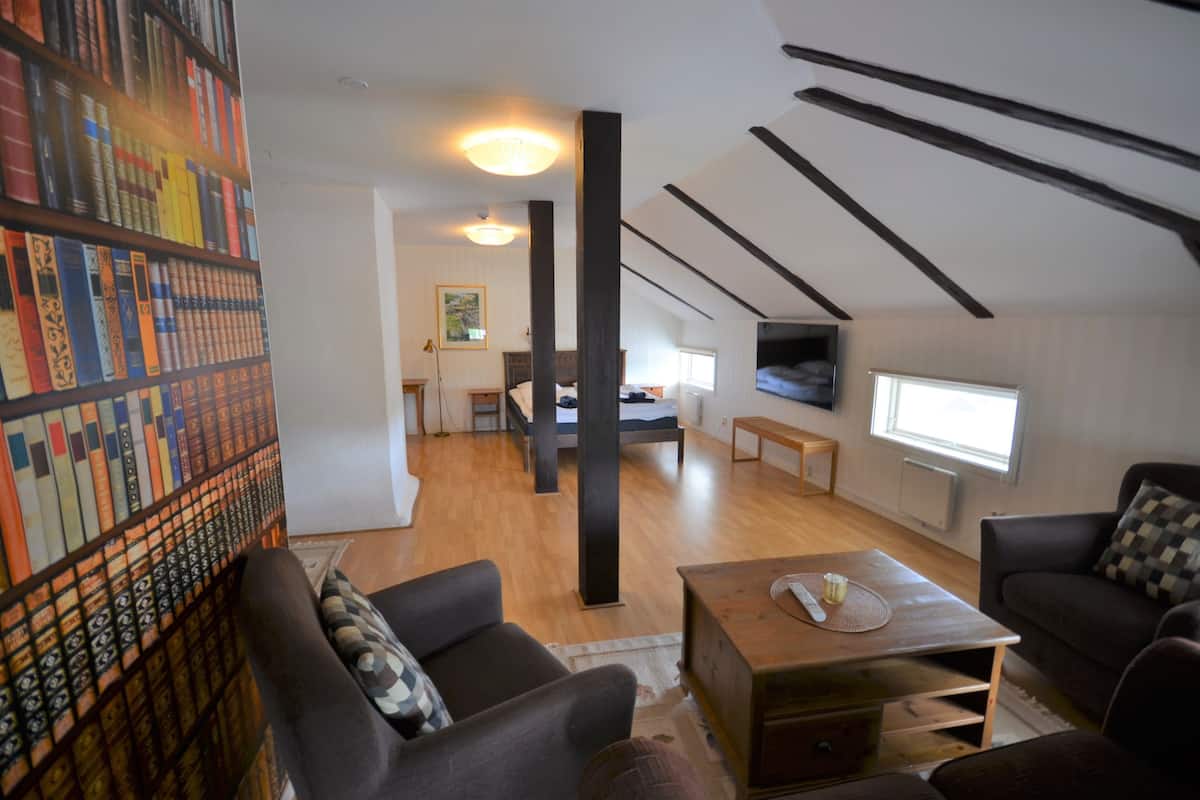 A spacious loft with high ceilings, exposed beams, a large bookshelf, a dining area, and a cozy seating area with a view of the city.