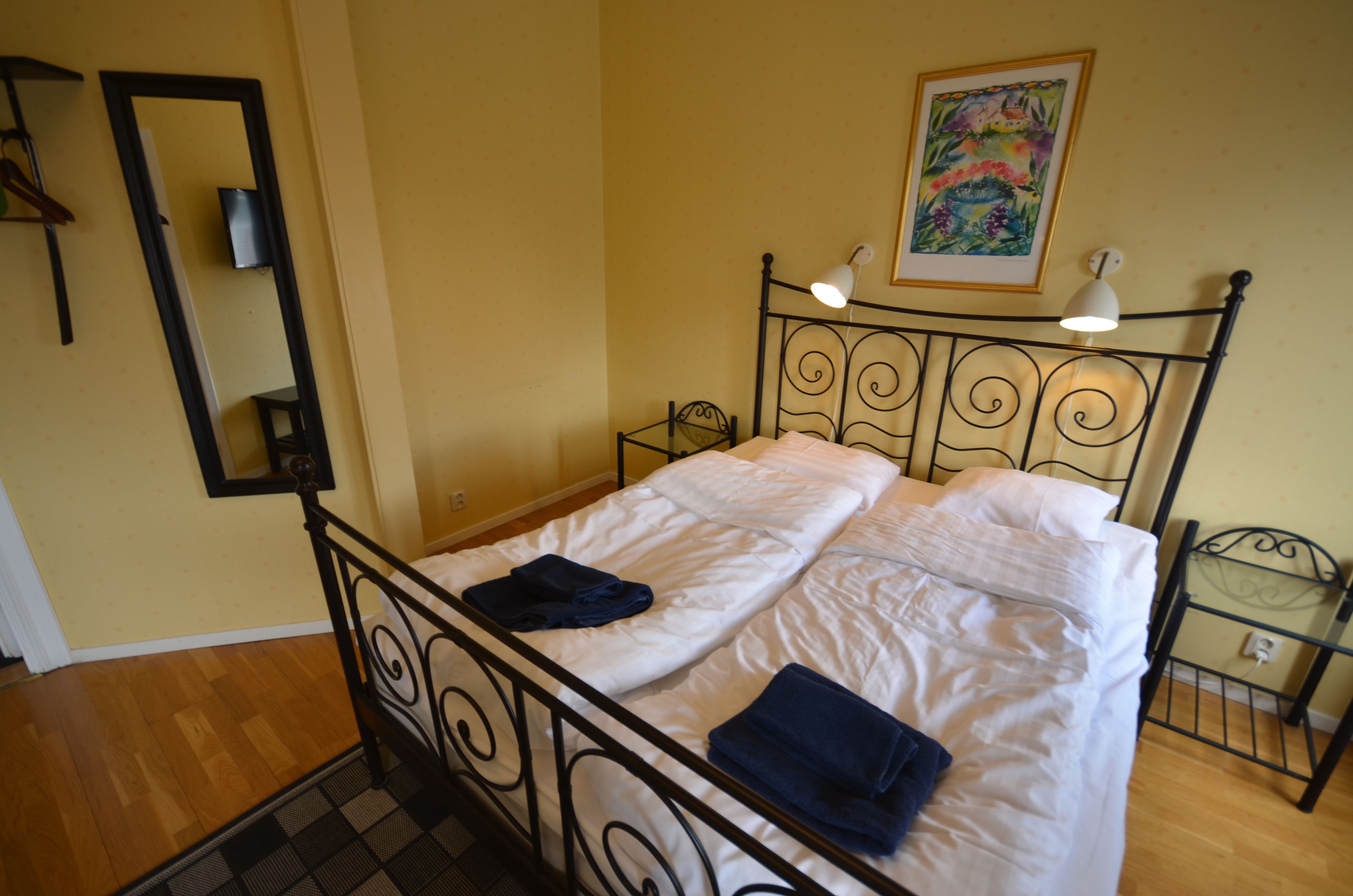 A neatly made bed with white linens and two pillows, a small bedside table with a lamp, and a painting on the wall above the bed.