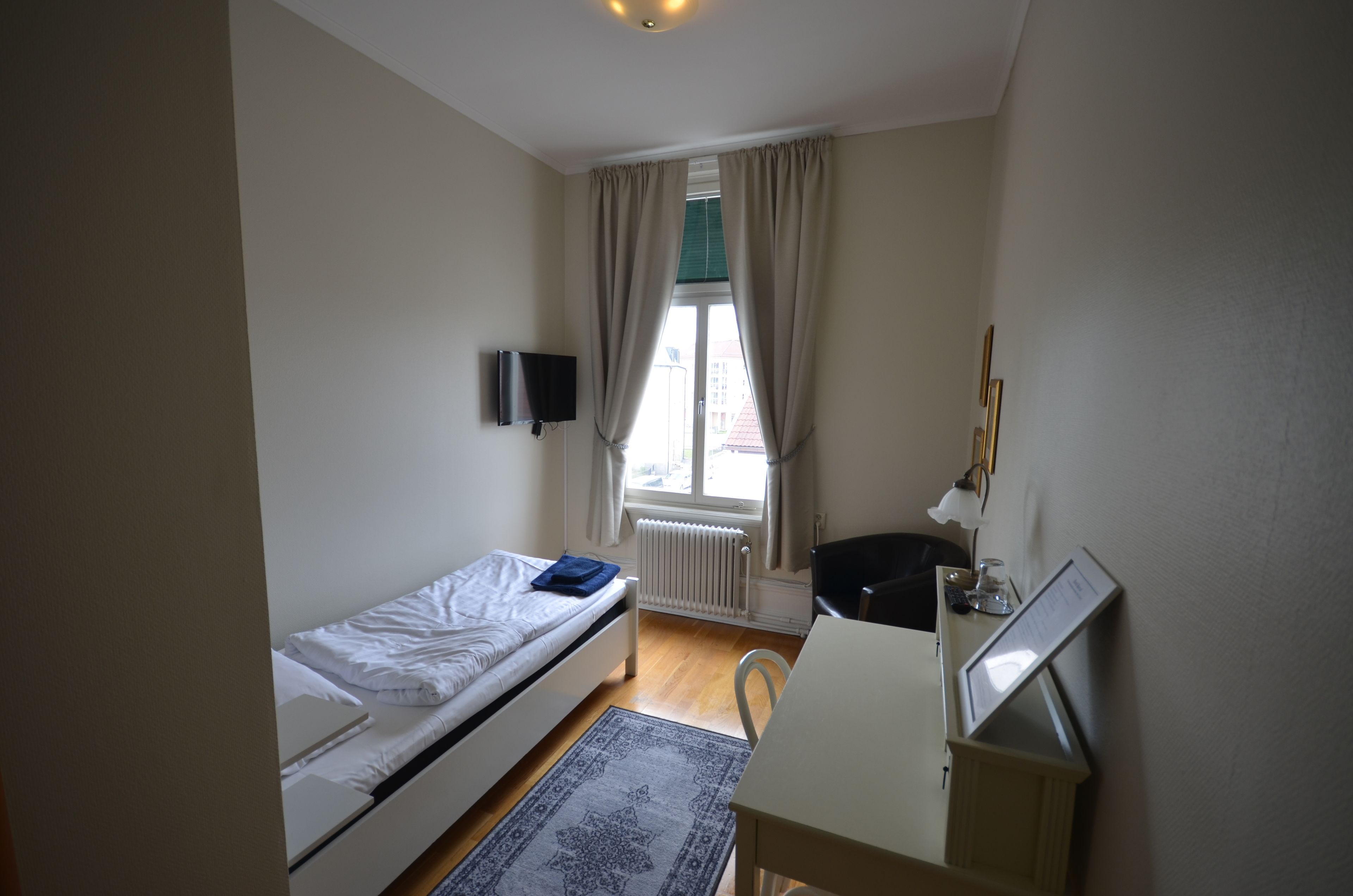 A small, well-lit room with a single bed, a desk with a chair, a radiator, and a window with curtains.