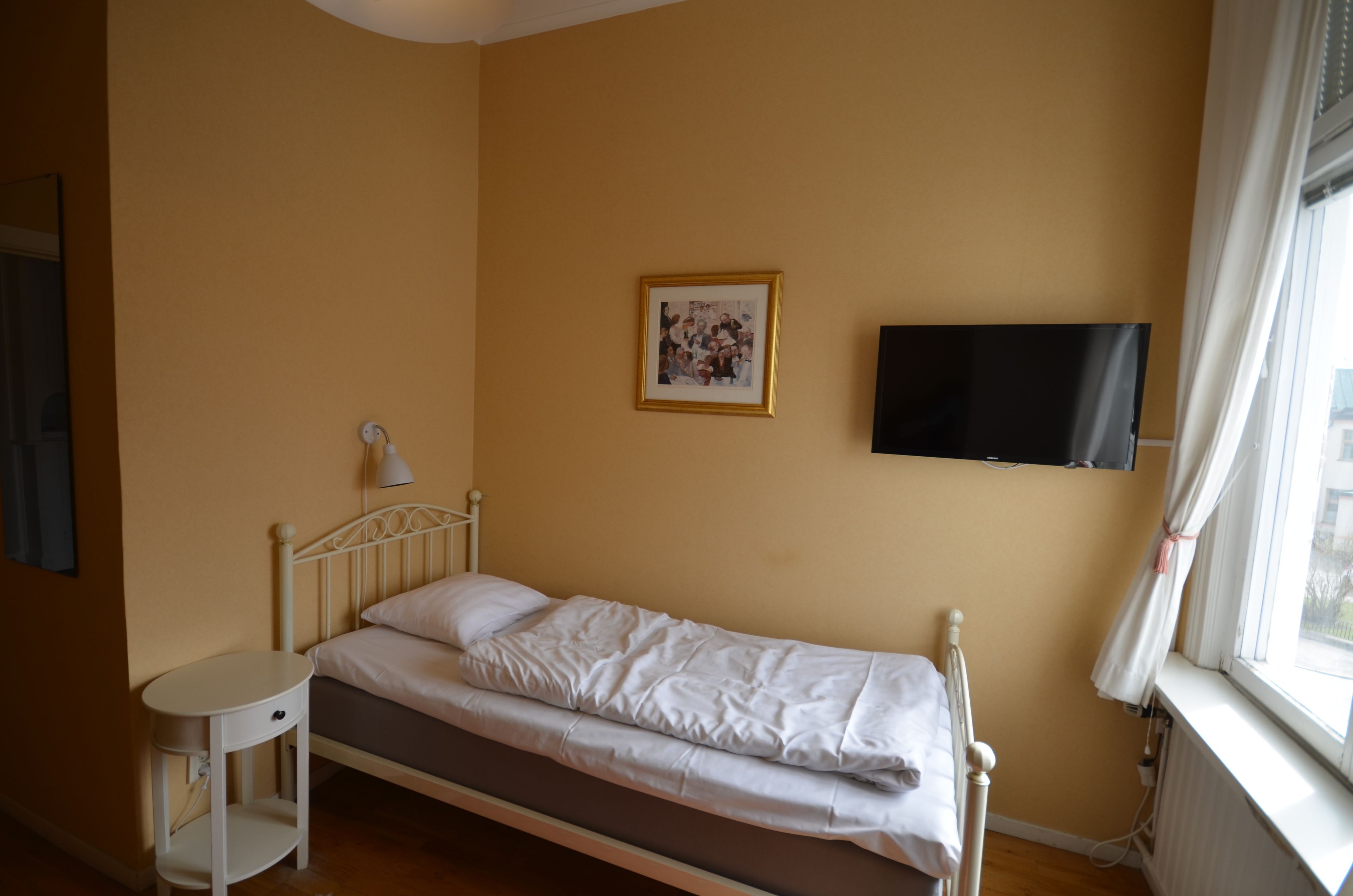 Un lit simple avec une tête de lit blanche, une table de chevet avec une lampe, une photo encadrée accrochée au mur et un téléviseur fixé au mur.