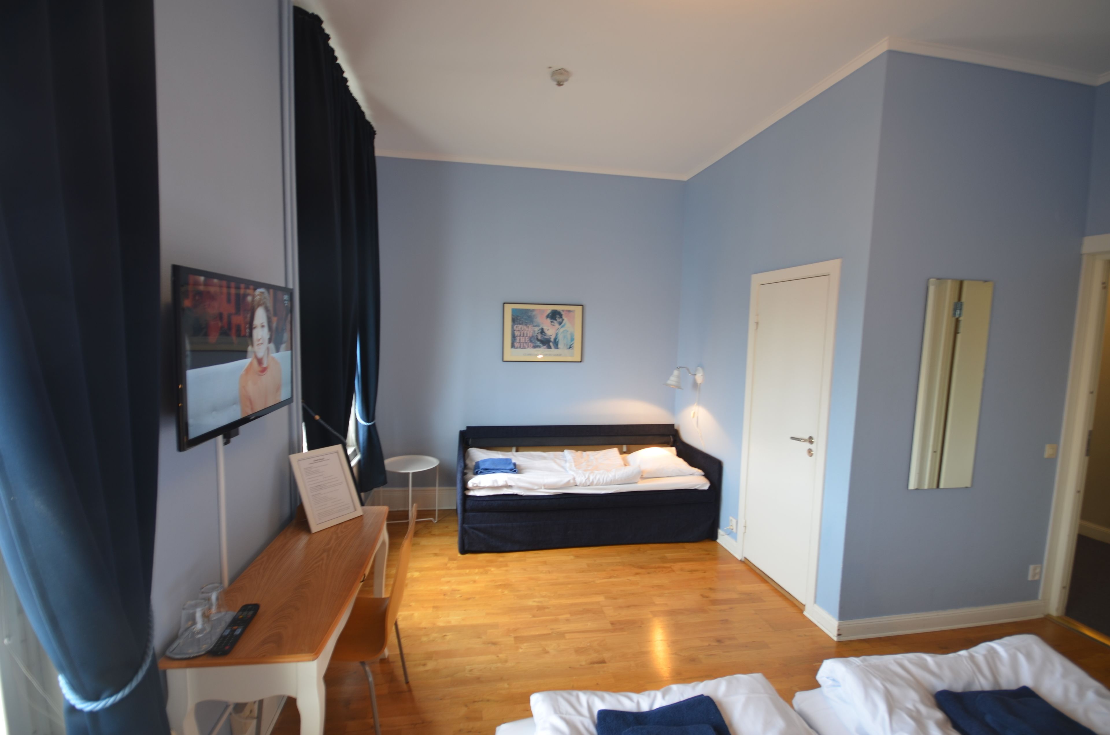 Una habitación con cama, escritorio, silla, una mesita y televisor en la pared.