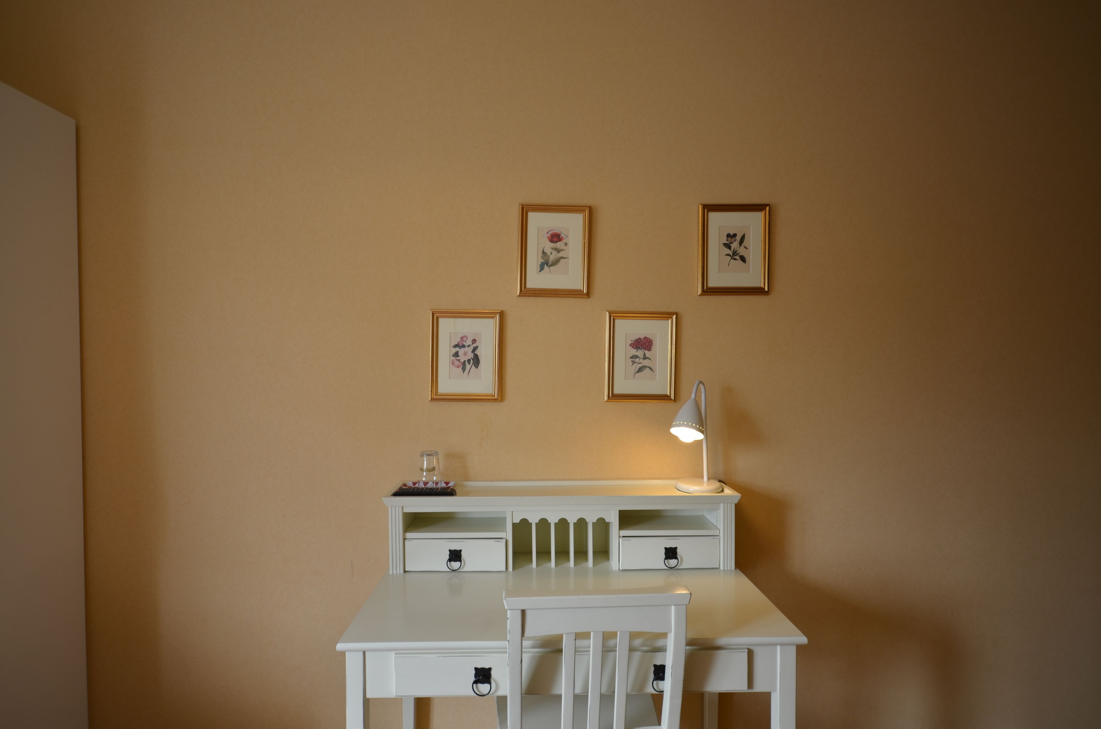 A white desk with a lamp, a chair, and four framed floral pictures on the wall.