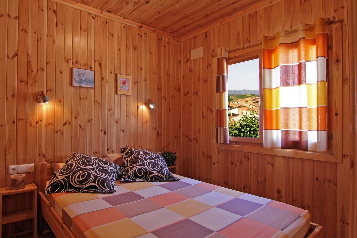 A wooden room with a bed, two wall-mounted lights, a window with curtains, and a small table with a plant.