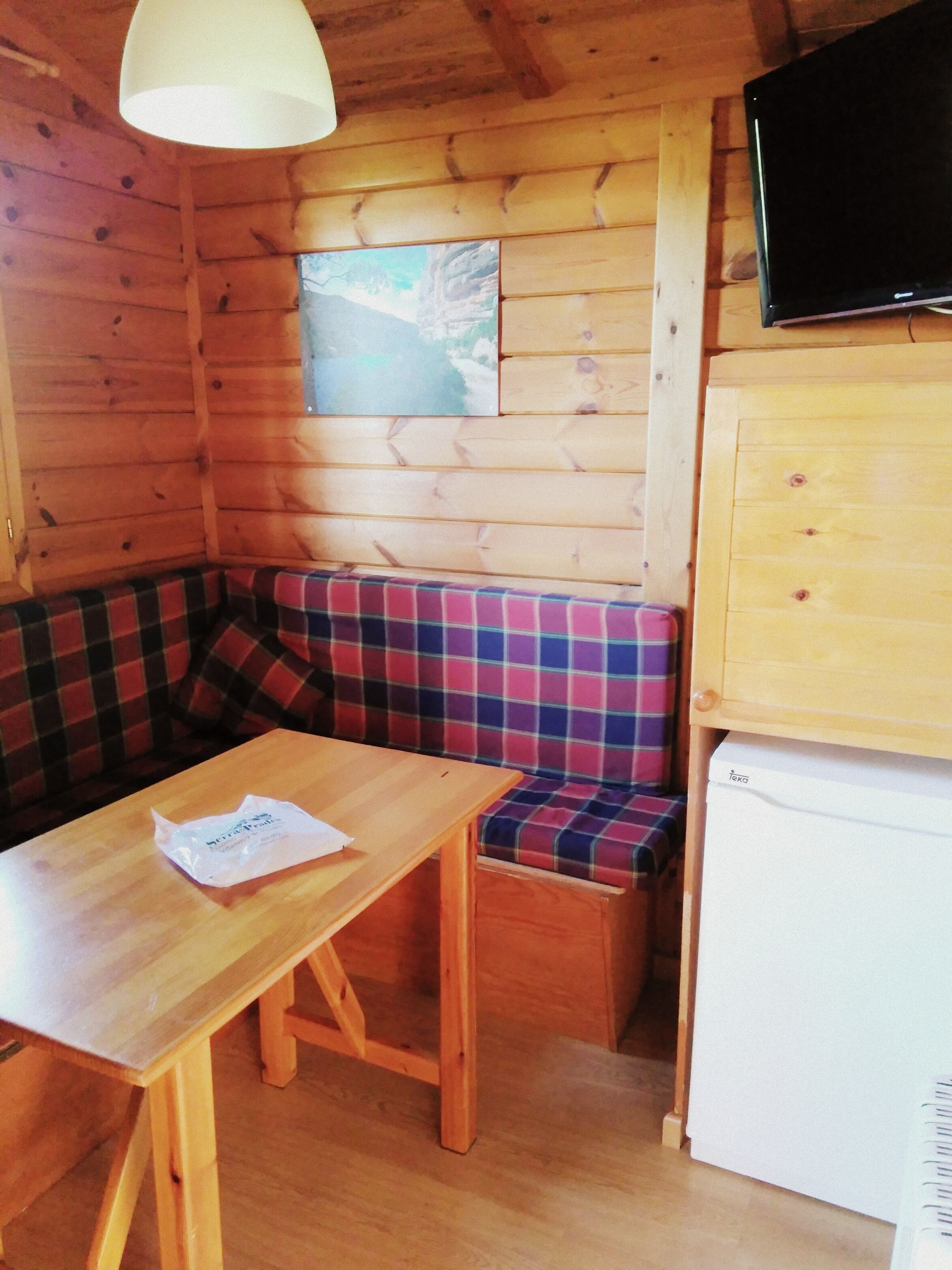 Interior d'una cabana de fusta amb taula de fusta, una banca estampada, televisió penjada i un frigorífic.