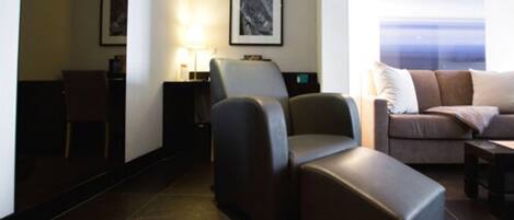 Habitación de hotel moderna con un sofá de tonos oscuros, un sillón reclinable y vista al mar a través de un ventanal grande.
