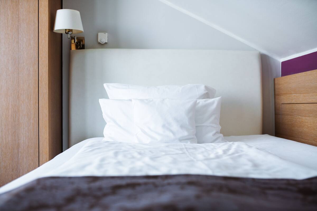 Een net opgemaakt bed met witte lakens en een donkerbruine plaid, een wandlamp en een houten hoofdbord.