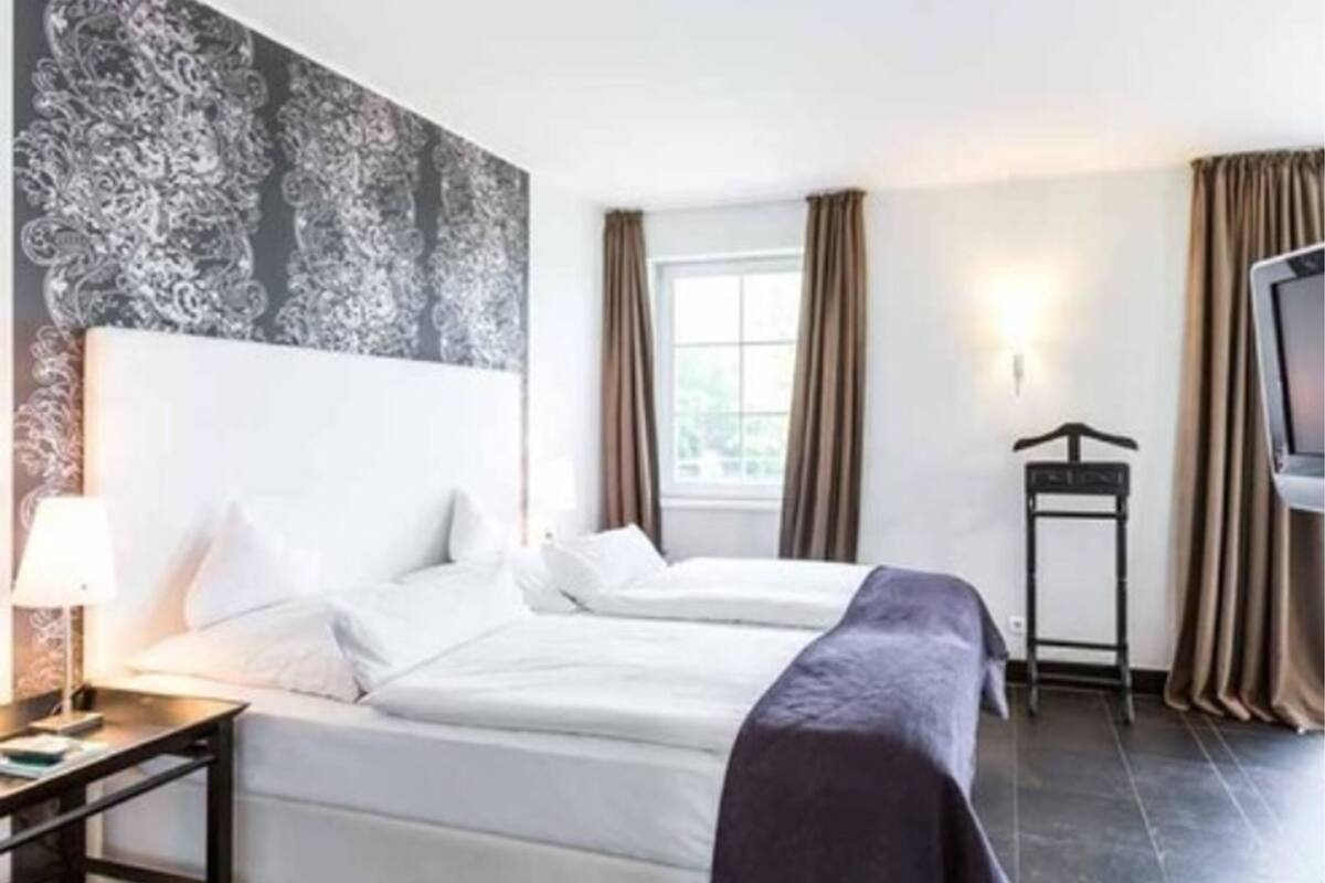 Een hotelkamer met twee bedden, een nachtkastje met een lamp, een tv-meubel en een raam met gordijnen.