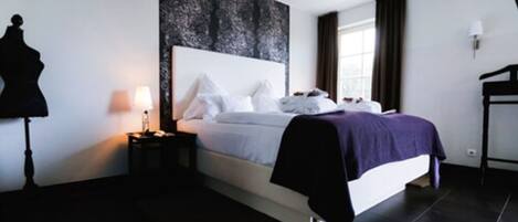 A modern hotel room with a large bed, a nightstand, a lamp, and a dark floor.