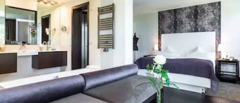 A modern hotel room with a large bed, a sofa, a desk, and a bathroom with a mirror and sink.