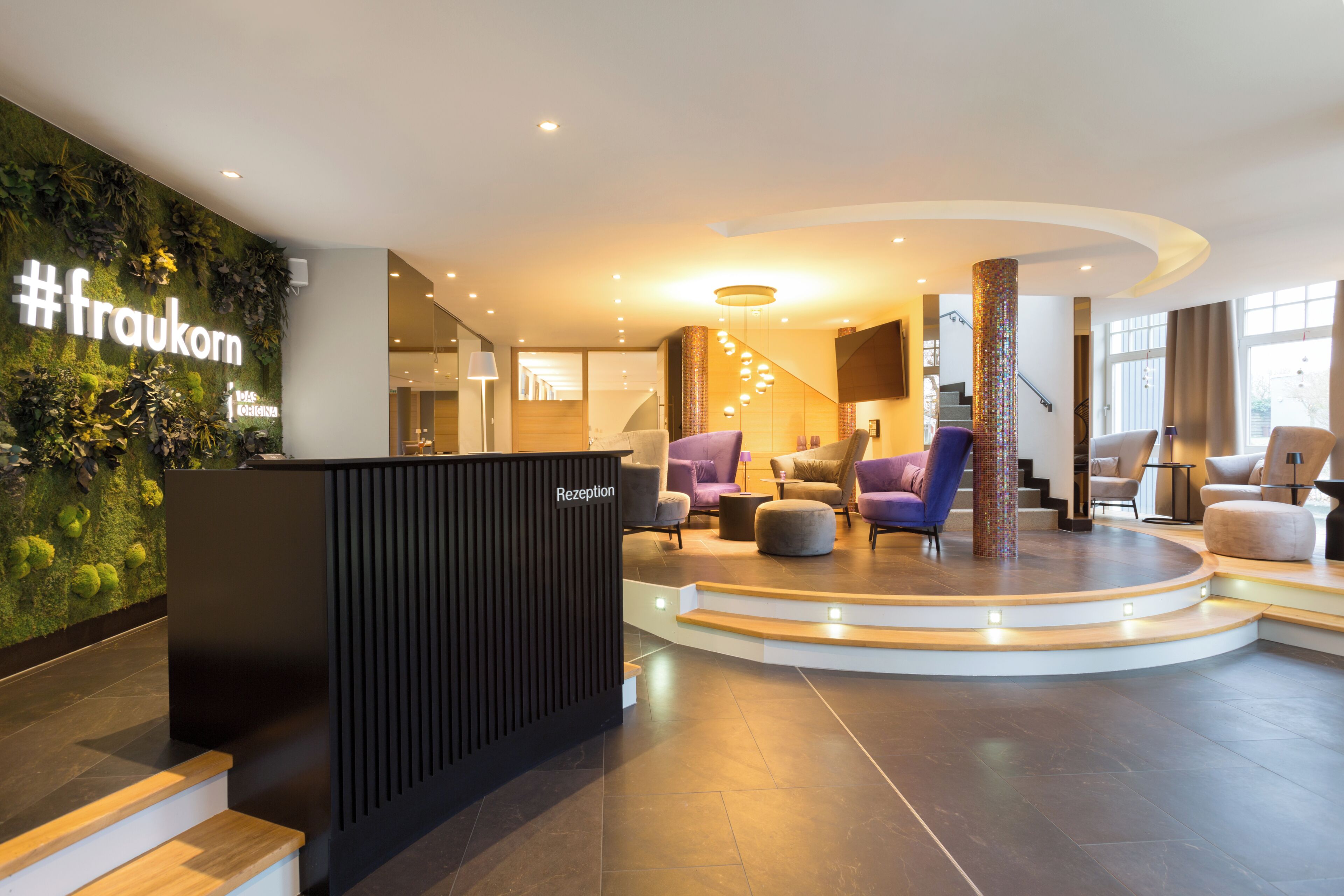 A reception area with a #fraukorn sign, a reception desk, a staircase, and a seating area with chairs and ottomans.