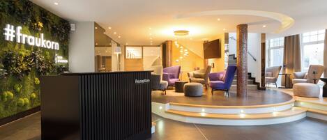 A reception area with a #fraukorn sign, a reception desk, a staircase, and a seating area with chairs and ottomans.