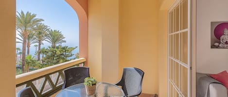 Ein Balkon mit Glastisch und Stühlen, einem kleinen Topfpflanzen und Blick auf Palmen und das Meer.