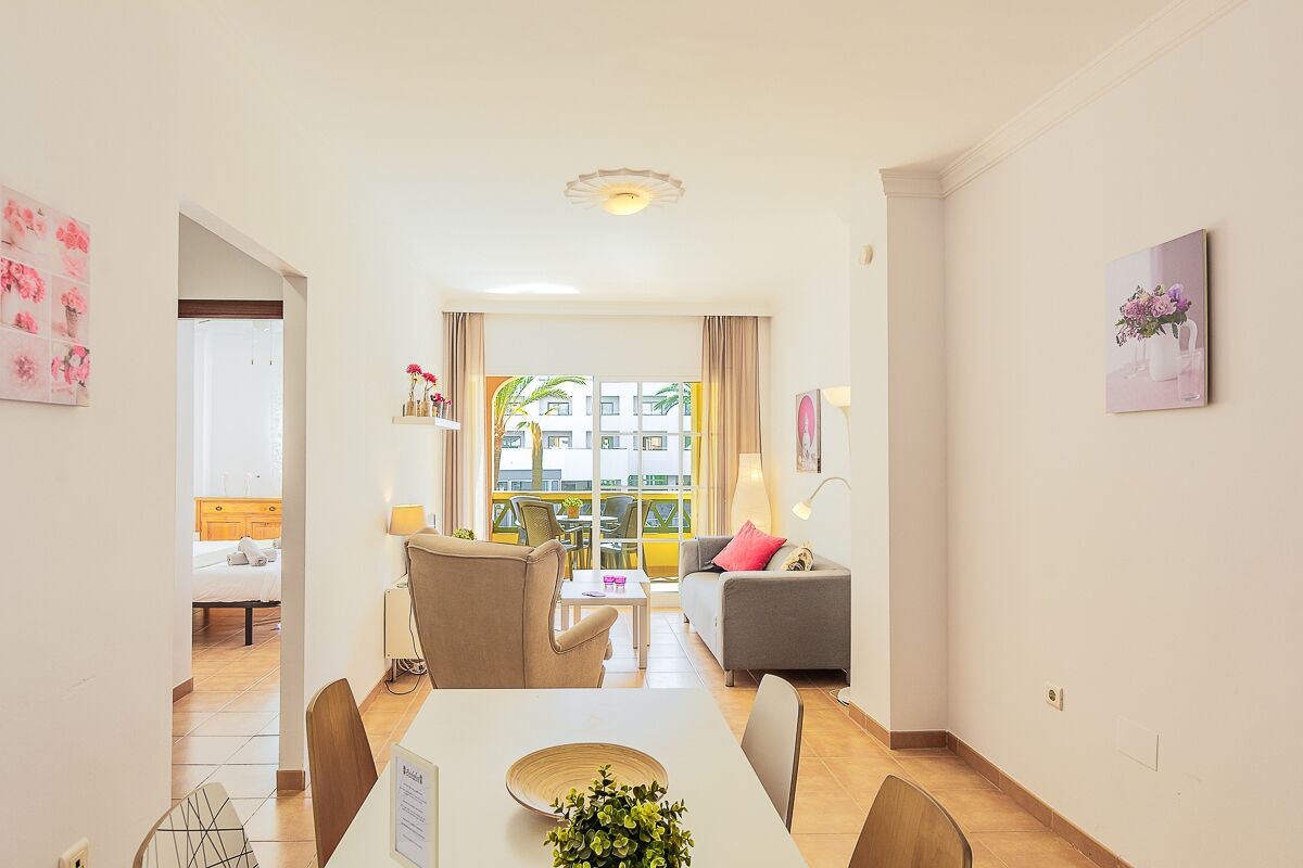 A modern living room with a dining area, a sofa, a coffee table, and a view through a large window.