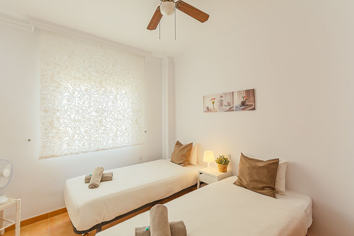 Une chambre d’hôtel avec deux lits, un ventilateur de plafond, une fenêtre avec des rideaux à motifs, une table de chevet avec une lampe et une plante, et une petite œuvre d’art accrochée au mur.