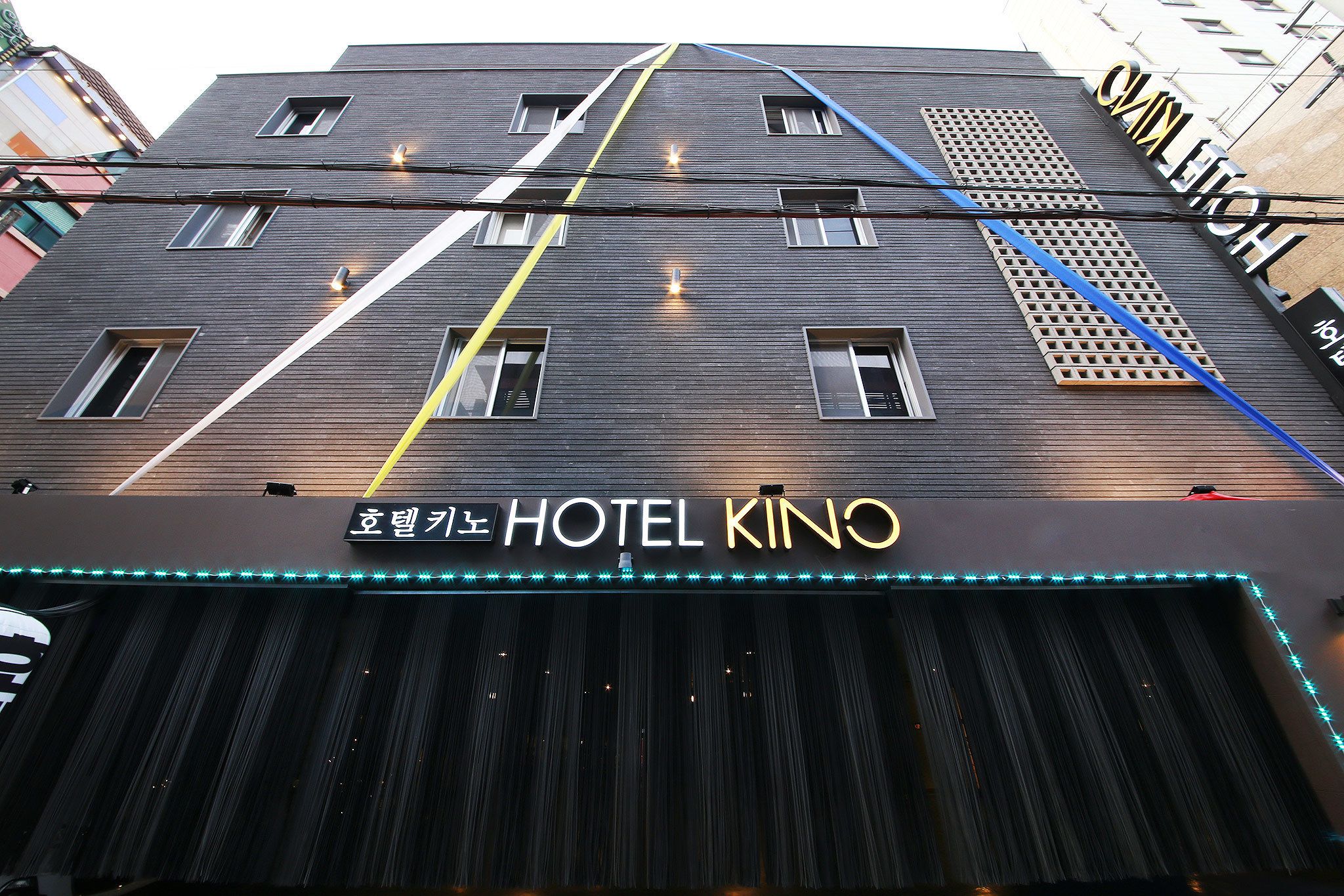 Um hotel com fachada moderna, um letreiro indicando "HOTEL KINO" e uma série de janelas verticais.