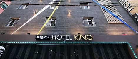 Um hotel com fachada moderna, um letreiro indicando "HOTEL KINO" e uma série de janelas verticais.