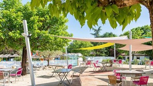 An outdoor seating area with tables and chairs under a shaded canopy, surrounded by trees and a pool. - Camping L'évasion - Mobilhomes (Puy-l'Eveque)
