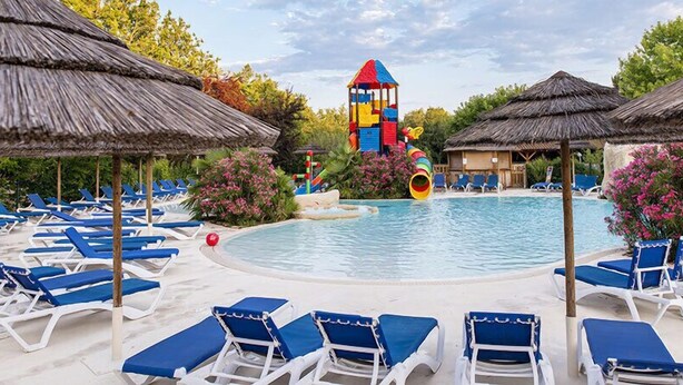 A pool area with blue lounge chairs, a colorful play structure, and thatched umbrellas. - Camping L'évasion - Mobilhomes (Puy-l'Eveque)