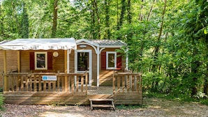 A wooden cabin with a porch, surrounded by dense forest. - Camping L'évasion - Mobilhomes (Puy-l'Eveque)