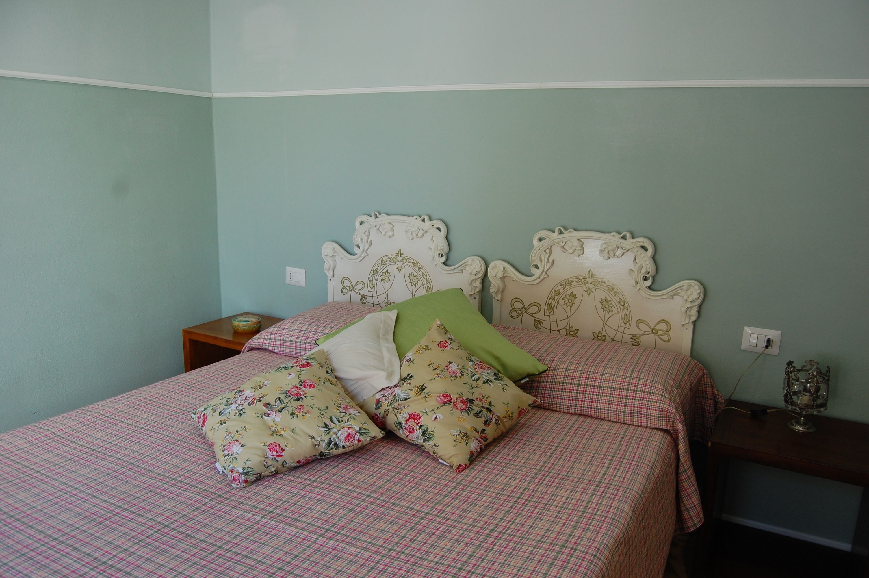 A bed with floral-patterned pillows and a checkered bedspread, a nightstand with a lamp, and a decorative wall piece.