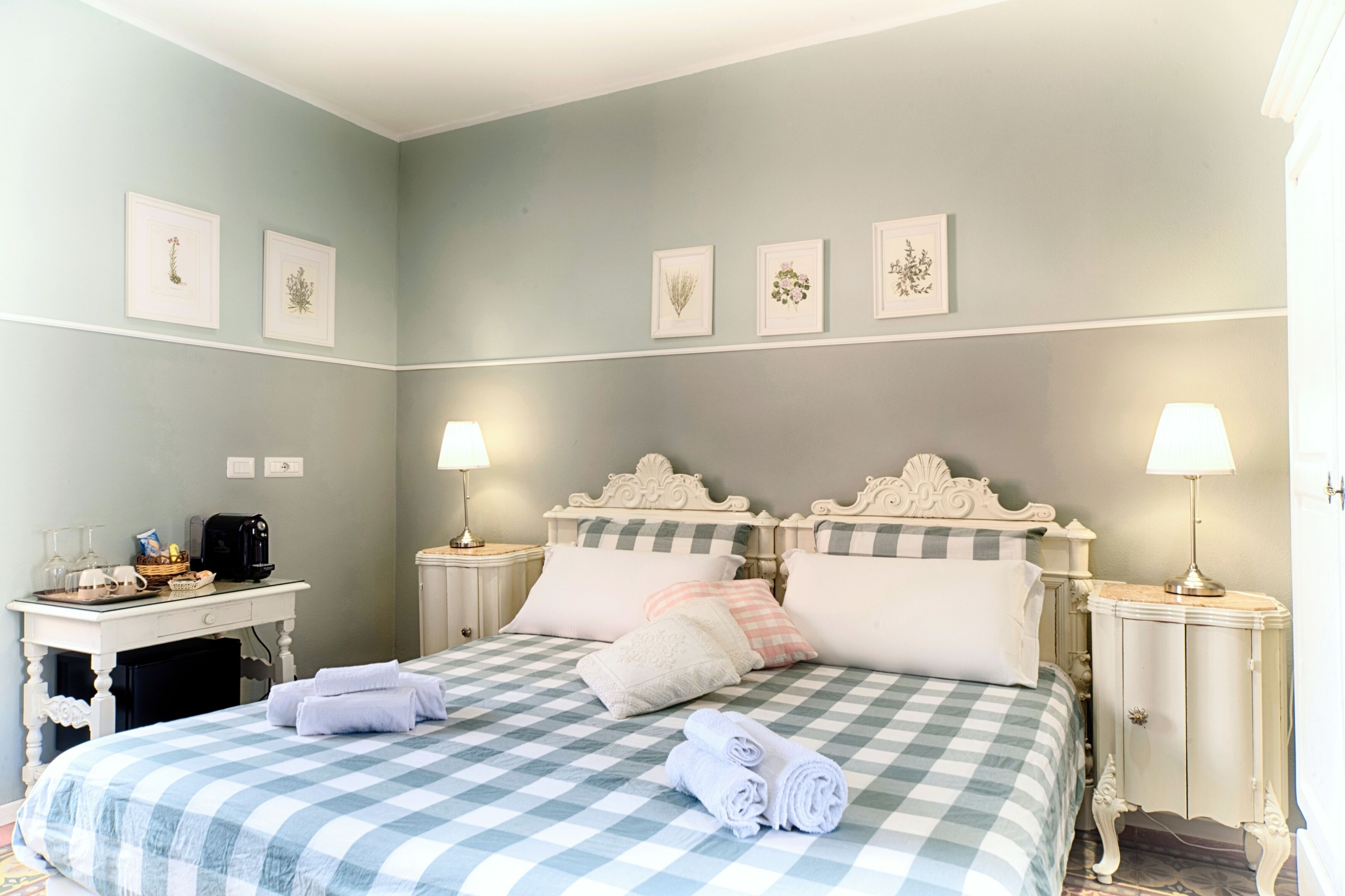 A neatly arranged bedroom with a checkered bedspread, two bedside tables with lamps, a desk with a coffee maker, and framed botanical prints on the wall.