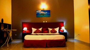 A hotel room with a large bed, two bedside lamps, a painting on the wall, and a small table with a chair. - Hotel Hilltop Country Club (Kigali)