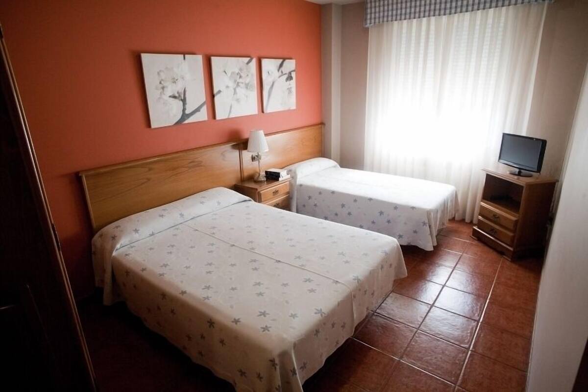 Habitación de hotel con dos camas, cabecera de madera, mesita de noche con lámpara, televisor sobre un mueble y una obra de arte en la pared.