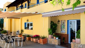 An outdoor cafe area with tables and chairs, potted plants, and a building entrance. - Hospedería Ancladero (Mojacar)