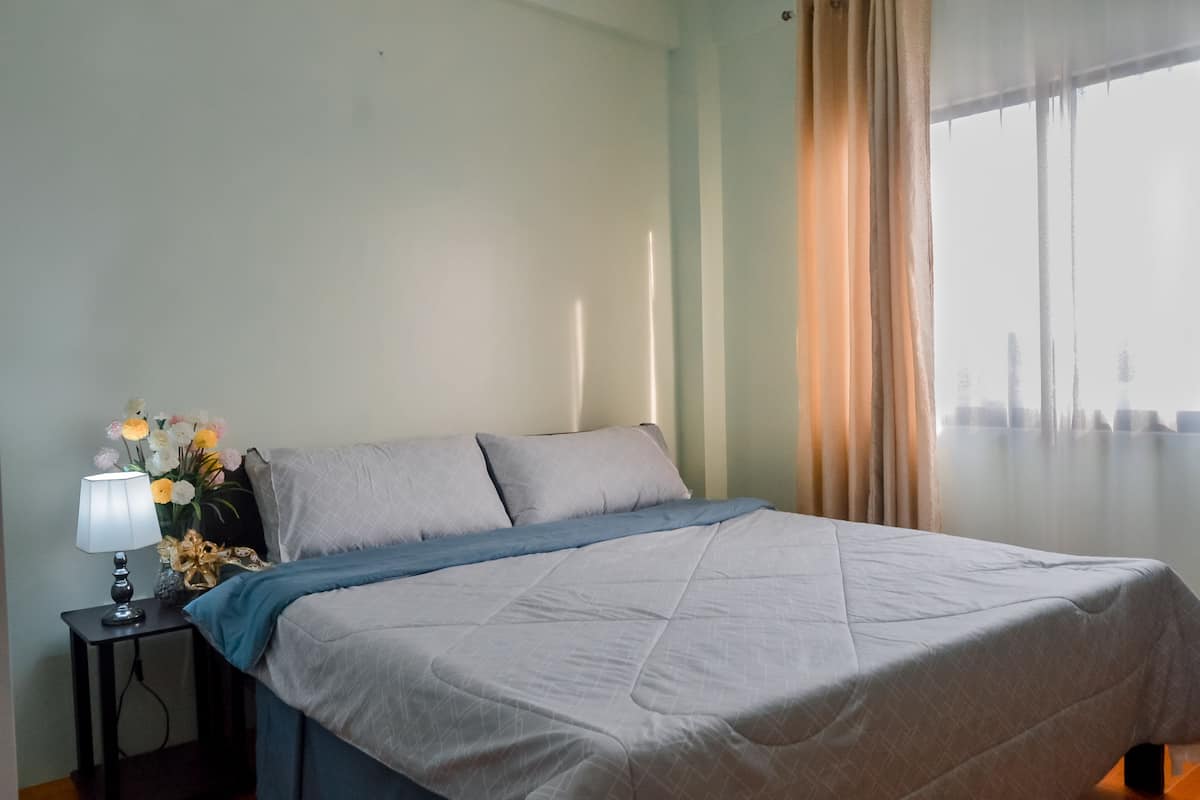 A neatly made bed with a blue blanket, a bedside table with a lamp and flowers, and a window with curtains.