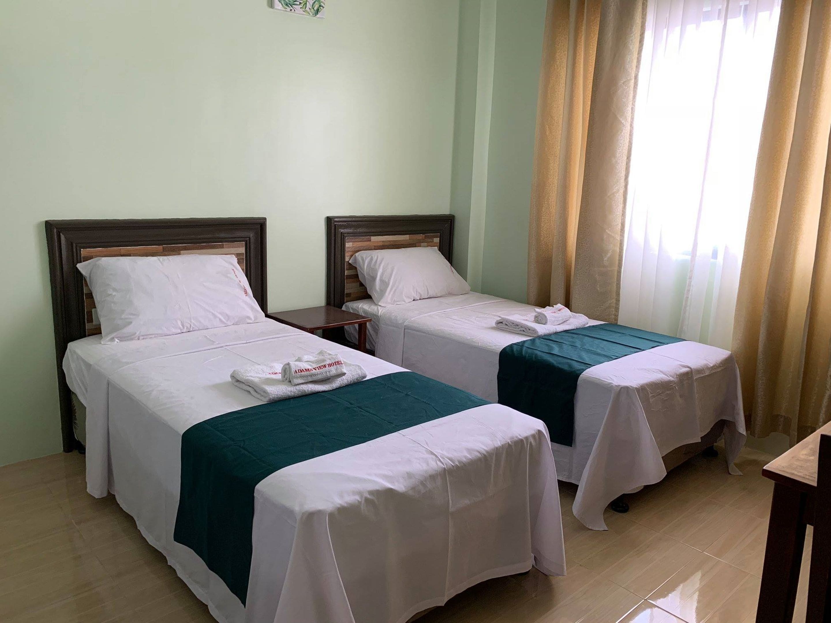 Une chambre d’hôtel avec deux lits simples, une tête de lit en bois, une petite table et une fenêtre avec des rideaux.