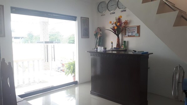 A reception area with a dark wooden desk, a clock, and a wall clock above it. There are clocks labeled 'USA', 'Spain', and 'Philippines'. A potted plant and a vase with flowers are on the desk. - Adams View Hotel (Moalboal)