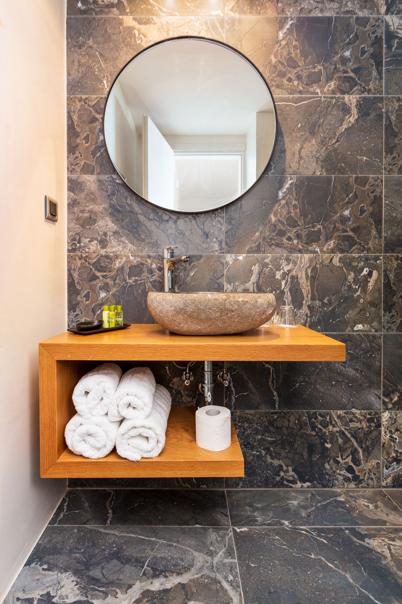 Een badkamer met een marmeren spatwand, een ronde spiegel, een houten wastafel en een plank met handdoeken en toiletartikelen.