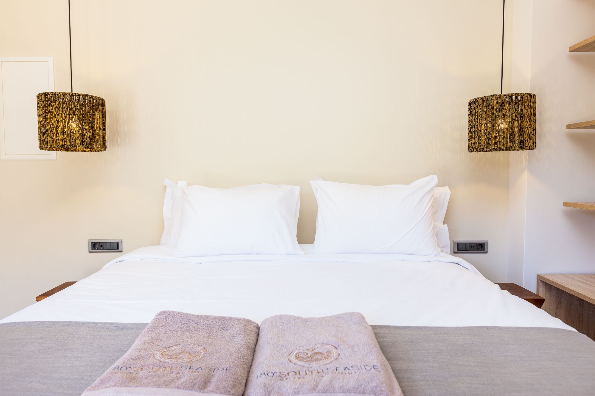 A hotel room with a bed, two white pillows, two towels with text, two hanging lamps, and a bedside table.