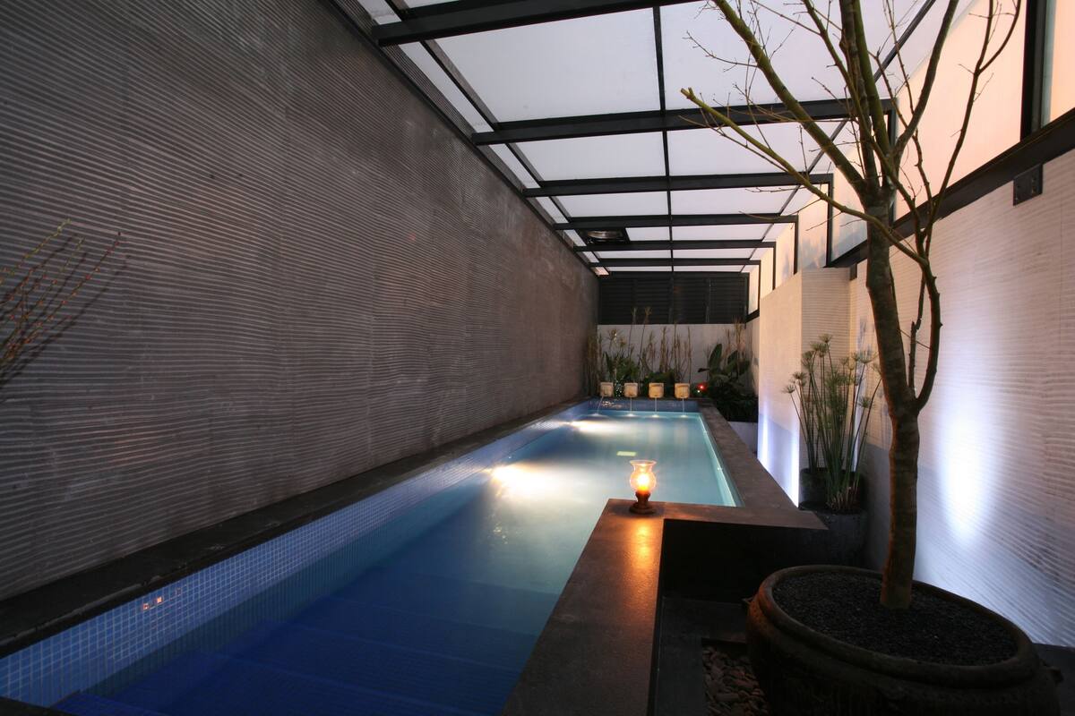 Una piscina interior con suelo de baldosas, techo de cristal y una pequeña mesa con una lámpara.