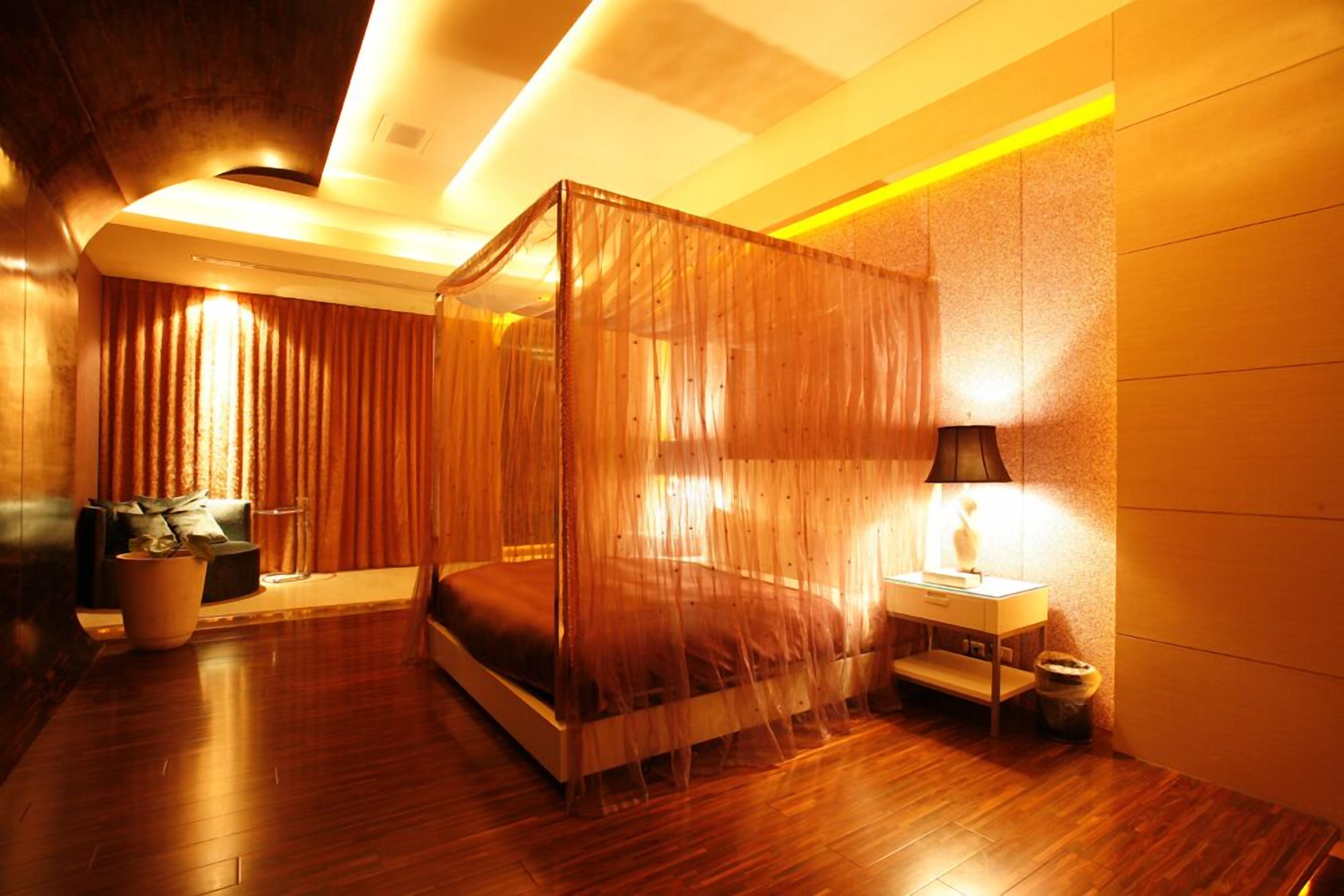 A modern bedroom with a four-poster bed, a bedside table with a lamp, and a potted plant.