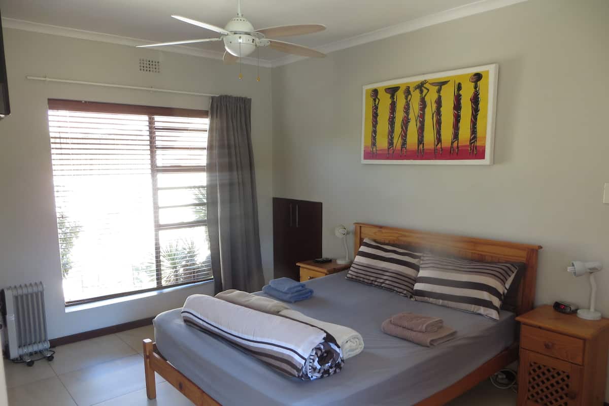 A bedroom with a bed, a ceiling fan, a window with curtains, a radiator, a nightstand, and a wall painting.