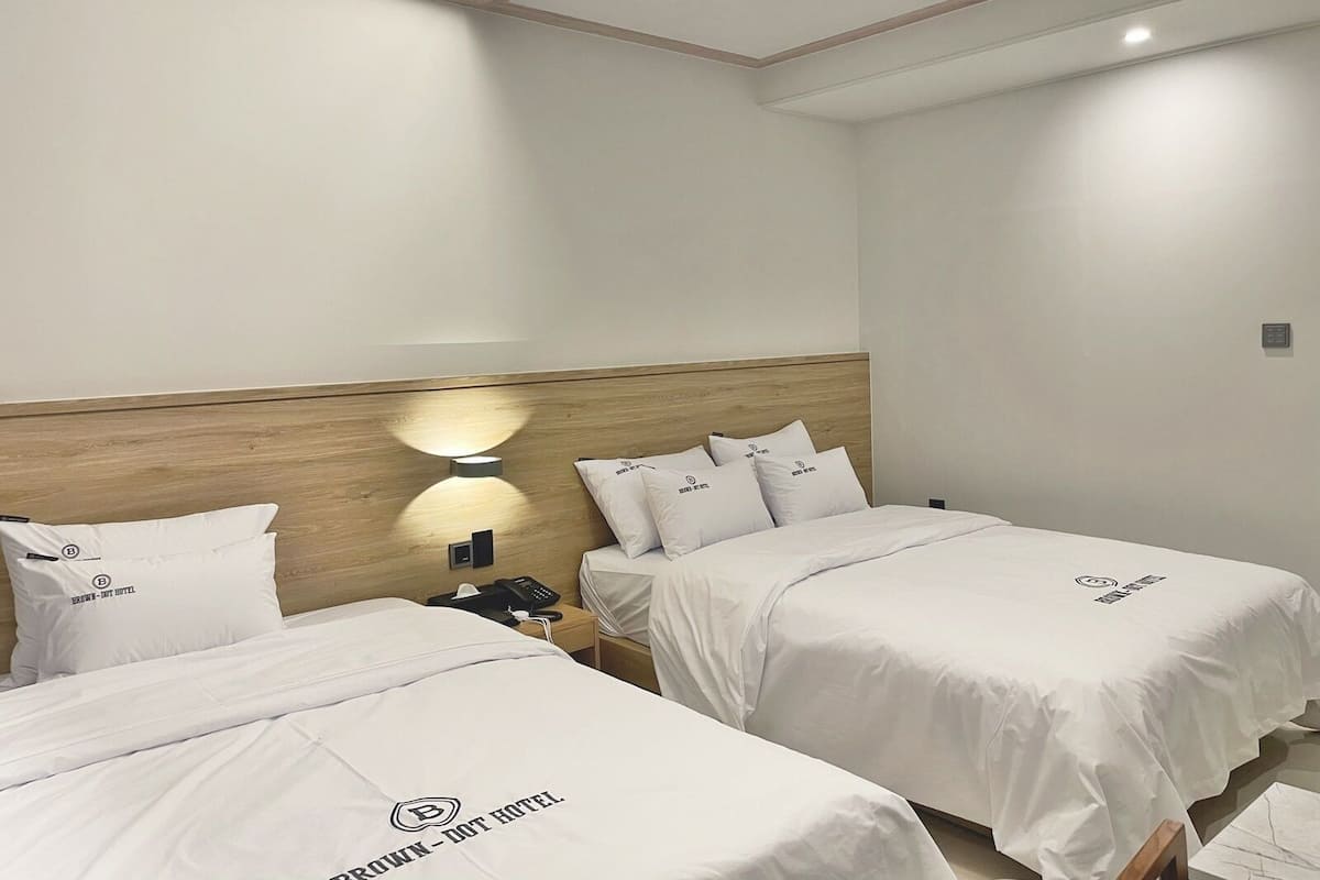 Ein Hotelzimmer mit zwei Betten, beide mit weißer Bettdecke mit dem Hotel-Logo. Dazwischen befindet sich ein Nachttisch mit Telefon und Lampe.