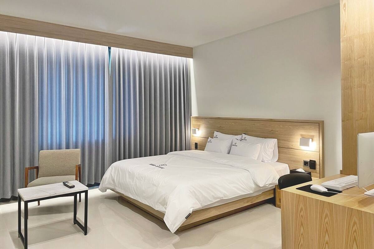 Ein modernes Hotelzimmer mit einem großen Bett, einem Schreibtisch mit Computer, einem Sessel und einem Fenster mit Vorhängen.
