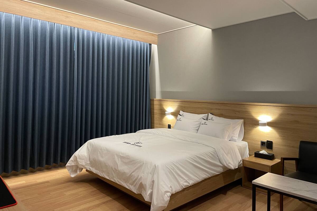 Ein modernes Hotelzimmer mit einem großen Bett, einem hölzernen Kopfteil, zwei Nachttischlampen, einem Nachttisch, einem Schreibtisch und einem großen Fenster mit blauen Vorhängen.